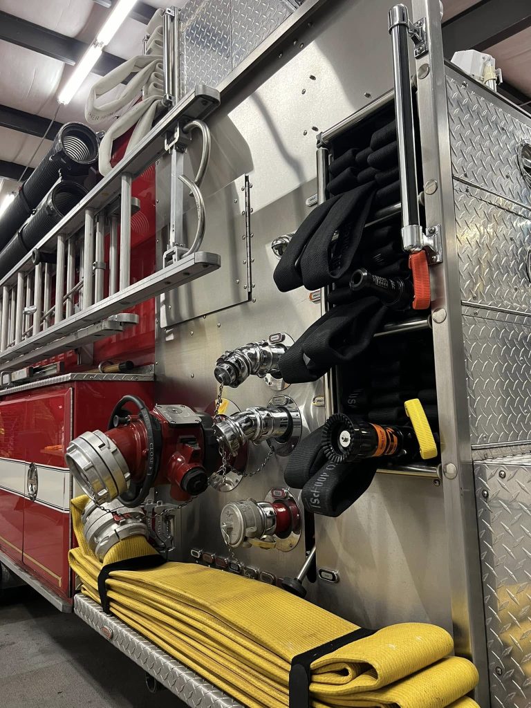 A close-up of a fire truck's side, showing various firefighting tools. Coiled hoses are neatly arranged next to nozzles and connectors. The vehicle features a shiny red and silver exterior, with additional equipment stored in compartments.