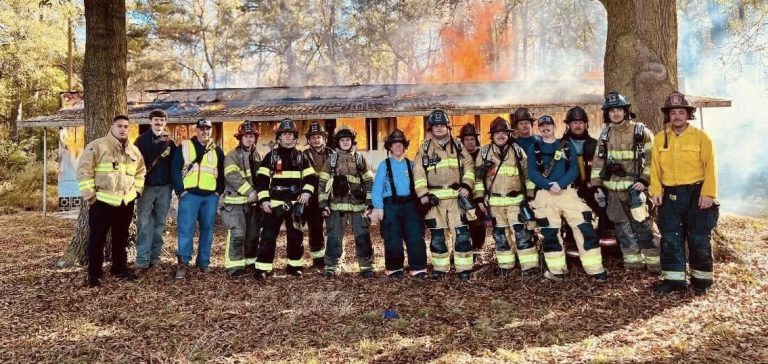 A group of firefighters and emergency personnel posing in front of a burning structure in a wooded area. They are wearing various protective gear and safety vests. Two large trees frame the scene, with smoke and flames visible in the background.