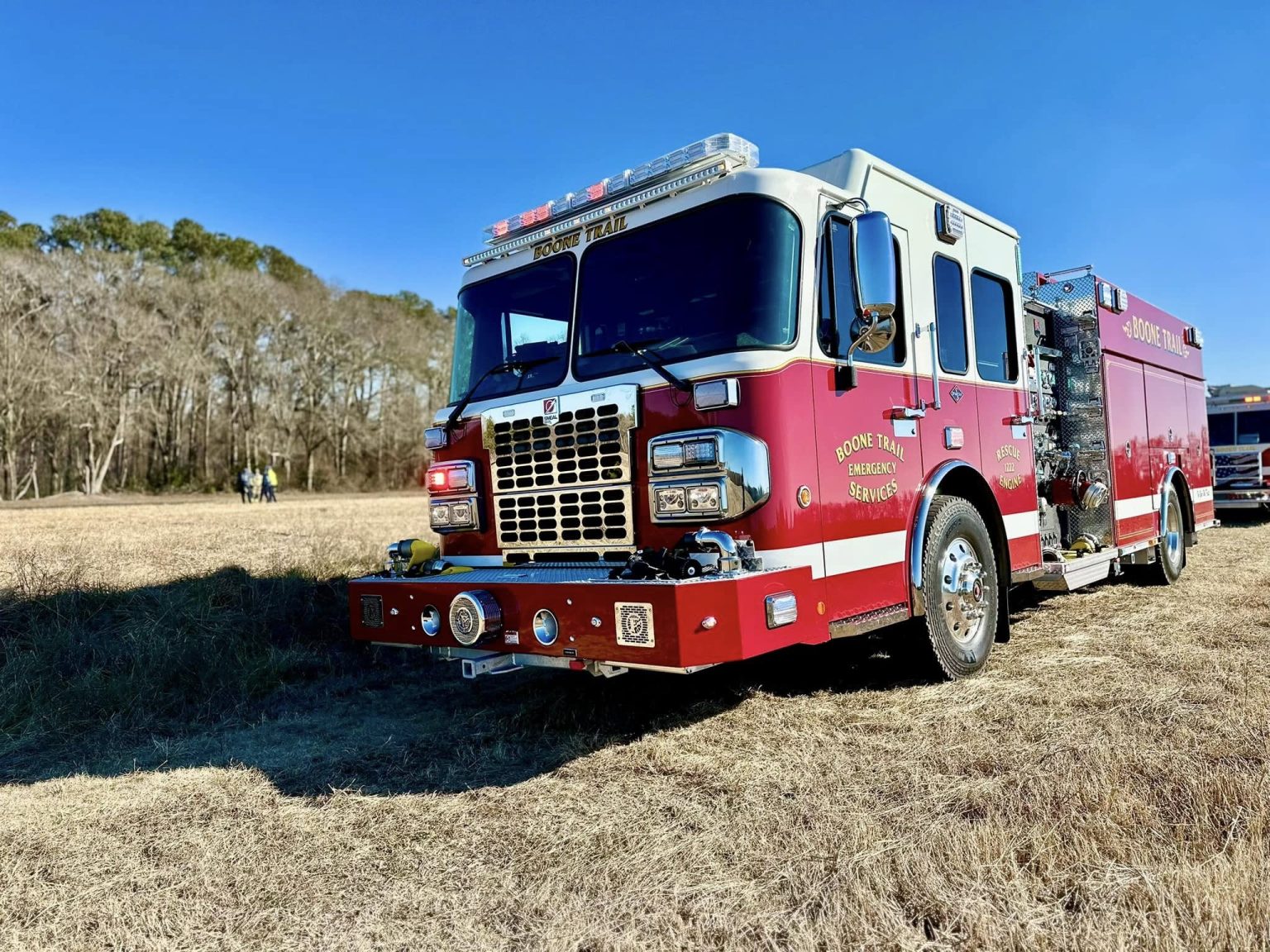 A red fire truck with "Hope Mills Fire Department" markings is parked in a grassy field under a clear blue sky. In the background, several people stand near a tree line.