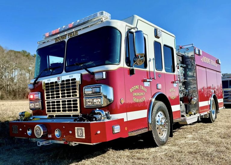 A red fire truck with "Hope Mills Fire Department" markings is parked in a grassy field under a clear blue sky. In the background, several people stand near a tree line.